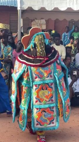 The video shows a person in a very elaborate, colorful costume with large padded shoulders and a tall, ornate headpiece. The costume is covered in intricate beadwork and embroidery featuring stylized animal figures and geometric patterns in shades of turquoise, orange, red, and yellow. The person is standing in an outdoor area with a dirt ground. In the background, there are spectators, some sitting and some standing, observing the person in the costume. The spectators appear to be of various ages and are dressed in casual clothing. There is a white tent structure visible in the background. The lighting suggests it is daytime.
