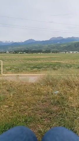 The video shows a person's legs in blue jeans in the foreground, with a vast, open landscape stretching out behind them. The scene is dominated by a wide, green field, likely a pasture or farmland, bordered by a fence with yellow posts and white rails. In the distance, rolling hills transition into majestic mountains, some of which are capped with snow, suggesting a cooler climate or high altitude. A small village or settlement is visible nestled amongst the hills, with trees and buildings dotting the landscape. The sky is overcast, indicating a cloudy day, but the light suggests it is daytime, possibly late morning or afternoon. The overall mood is serene and tranquil, with a sense of openness and connection to nature. The location context places this scene in Dolan, Kazakhstan, a region known for its scenic natural beauty and mountainous terrain. There is no visible text in the media file.