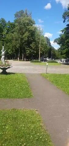 A slow pan right reveals a paved path in the foreground, bordered by a grassy area. Beyond that, a circular paved area with parked cars is visible, surrounded by trees. Further to the right, a swing set with people on it is situated under a canopy. The camera continues to pan right, showing more of the grassy area and trees. In the distance, a playground with a colorful seesaw and slide is visible. The sky is blue with scattered clouds.
