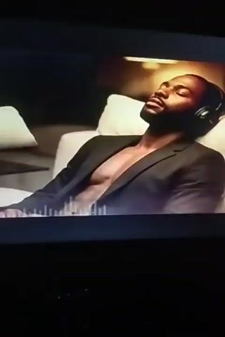 A man with his eyes closed and wearing headphones rests on a white couch. He is wearing a black blazer, open at the chest, revealing his torso. A portion of an equalizer is visible at the bottom of the frame. The background is dark, with a lamp visible in the upper right corner.