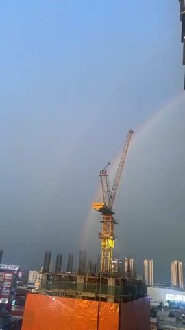 The video captures a construction crane silhouetted against a dusky, pale blue sky. A faint double rainbow arches across the upper right portion of the frame. The crane, predominantly yellow, is positioned in the center, its boom extending diagonally upwards and to the right. At the base of the crane, an unfinished building structure is visible, covered in orange protective sheeting. The building features exposed rebar protruding from concrete columns. In the background, the faint outlines of other buildings and city lights can be discerned. The lighting suggests either dawn or dusk, with a soft, diffused light illuminating the scene. No people are clearly visible. The camera appears to be positioned at a high vantage point, likely from a window or another elevated structure, looking towards the construction site. The video is short, a mere few seconds, and seems to be a static shot with no camera movement.