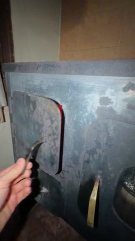 The video shows a close-up of a wood-burning stove with the door slightly ajar. Inside the stove, embers glow a bright red color amongst some partially burnt pieces of wood and ash. The interior walls of the stove are lined with brick. The stove is made of a dark metal and appears to be aged. The background is partially visible and suggests the stove is located indoors.