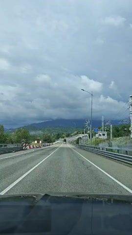 A car drives on a bridge on a two-lane road under a cloudy sky. A truck is driving in the same direction ahead of the car. Mountains in the background are partially obscured by the clouds. The road is lined with streetlights and guardrails. Construction cones are seen on the left side of the road.