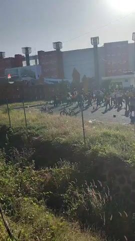 A group of people are gathered outside a building. Some are standing and watching, while others appear to be exercising or dancing. Several bicycles are parked nearby. The sun is shining brightly, casting long shadows. The overall atmosphere seems energetic and positive.