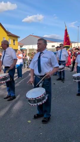 A group of people in matching uniforms, some playing drums, are marching in a parade. The central figure in the foreground is a man with graying hair, wearing a white shirt, blue tie, and blue pants, holding drumsticks and a snare drum. He is looking forward, seemingly focused on playing. Behind him, other individuals are also playing drums, and a few are carrying flags. The participants are on a paved street, with buildings and trees visible in the background. The sky is blue with some clouds, suggesting it is daytime. The overall atmosphere appears to be a celebratory event or a marching band performance.