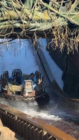 A raft-like amusement park ride vehicle moves through a waterway. The vehicle has multiple rows of seats, some of which are occupied by people wearing blue and yellow rain ponchos. The vehicle is surrounded by a rough, rocky environment with some vegetation overhead. Water splashes around the vehicle as it navigates the course, creating a dynamic and wet experience for the riders. The ride appears to be an outdoor attraction within a theme park.