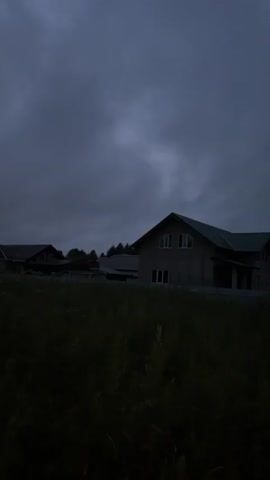 A static, wide shot captures a rural landscape at dusk. The foreground is dominated by dark, shadowy tall grass, obscuring much of the lower portion of the frame. In the mid-ground, several houses are visible, their roofs and structures silhouetted against the darkening sky. The most prominent house, in the center, appears to be under construction or unfinished, with visible siding. Behind the houses, trees form a darker, more dense line. The sky above is overcast, with heavy, dark clouds contributing to the dim, twilight atmosphere. The overall impression is one of quietude and the transition from day to night.
