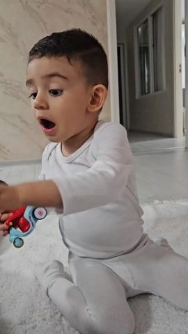 A small boy, likely a toddler, dressed in white long-sleeved pajamas, is sitting on a white, plush rug in a well-lit room. He is holding a toy that appears to be Mickey Mouse, with a red body and blue base. The boy first points with his index finger, then expresses surprise with an open mouth and wide eyes as he looks at the toy. He then looks to his right with an expression of shock or concern, possibly calling out "Papa." He then looks down at the toy with a more thoughtful expression. The background shows a hallway with a door and window, and the walls are a light neutral color.