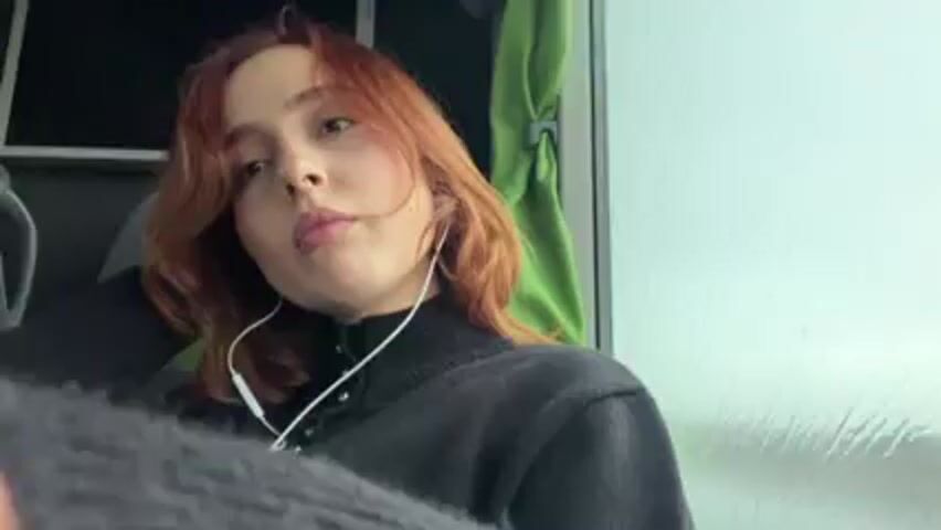 A person with red hair, wearing a black turtleneck sweater and white earphones, is looking to their left and touching their hair. They are sitting in a bus or train. The walls are white and there is a green curtain behind them. The camera is held by a person wearing a gray sweater, who is facing the red-haired person.