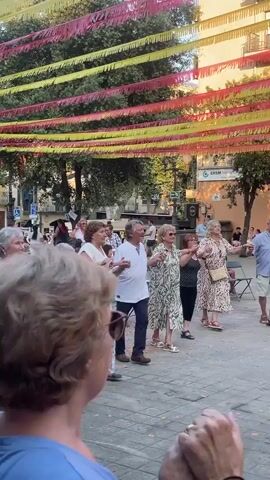 A group of older adults are dancing in a circle in an outdoor plaza. They are holding hands and moving in a synchronized fashion. The plaza is paved with cobblestones and decorated with colorful streamers hanging overhead. There are trees and buildings in the background. Some people are standing and watching the dancers. The dancers are dressed in casual summer clothing. The atmosphere appears festive and joyful.