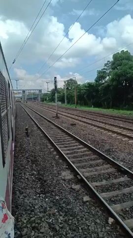 The video shows a view from inside a moving train, looking out the side and down the railway tracks. The train is traveling on the right side of the frame. There are multiple sets of railway tracks visible, with gravel ballast and ties. Overhead power lines with supporting poles are present. On the left side of the frame, the exterior of the train is visible, including windows and a door handle. In the distance, a railway station with a bridge-like structure above the tracks can be seen. Trees and vegetation line the right side of the tracks. The sky is cloudy. There are no people clearly visible in the frame.