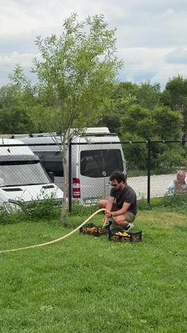 A man wearing a dark t-shirt and khaki shorts is crouching on a grassy lawn next to a small tree. He is holding a garden hose and directing water into two black plastic crates filled with yellow objects, possibly fruit. Behind him, two white vans are parked side-by-side, and a black fence runs along the edge of the property. In the background, lush green trees and vegetation are visible. A person is sitting to the right of the frame, partially obscured by the fence. The sky above is overcast with clouds.