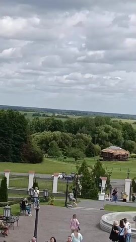 This is a video of a scenic overlook with people and buildings. In the foreground, there is a circular fountain with lion head spouts. Several people are standing around the fountain. To the left of the fountain, there is a white railing with decorative white pillars, and behind the railing, there are benches and manicured bushes. Further back, there are several buildings with brown roofs, surrounded by green fields and trees. In the distance, there is a body of water and a wide expanse of fields under a cloudy sky. Several tour buses and cars are parked in an area to the left of the buildings. People are walking around the grounds and taking pictures. The overall impression is of a well-maintained tourist attraction.