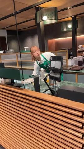 In a brightly lit café, a young woman with reddish-brown hair tied in a ponytail and wearing a white long-sleeved shirt and green apron is preparing a drink behind a marble-topped counter. She is focused on a tall mixing cup with a straw, which she then places on a digital scale before moving it. To her right, a black tablet is mounted on an adjustable stand. Another person in a yellow shirt is visible in the background, also working behind the counter. In the foreground, there's a wooden slatted counter. The café has a modern design with dark green and wooden accents.
