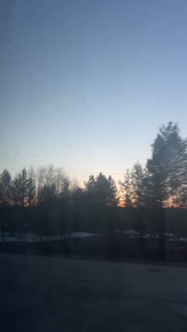 The video, taken from inside a moving vehicle, shows a landscape at dusk. A line of trees, mostly bare, forms the horizon against a sky with a gradient of blue to orange, indicating a sunset or sunrise. Patches of snow are visible on the ground. Utility poles and wires appear in the foreground as the vehicle progresses. The camera's movement causes blurring, particularly with closer objects. No people are visible.