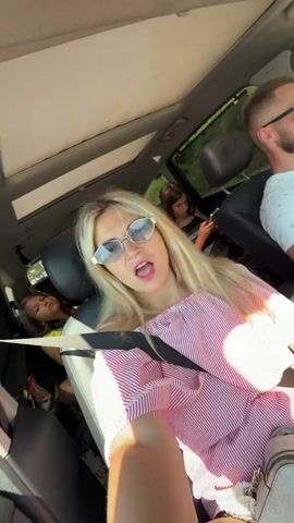 This is a first-person perspective video taken from inside a car. The camera person, a woman with blonde hair wearing sunglasses and a pink and white striped off-the-shoulder top, is in the passenger seat. A man, also wearing sunglasses, is driving the car. In the back seat, two young girls are visible. The car appears to be moving, as indicated by the blurred background and the slight motion of the vehicle. The sun is shining, creating bright light.
