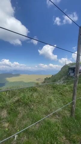 The video shows a scenic view from a mountaintop. In the foreground, there is green grass with some small yellow flowers and a wire fence. The background features rolling hills and mountains under a blue sky with white clouds. At one point, a person wearing a black cap and sunglasses is shown close-up, looking nervous and biting their nails. There are also some objects on a pole, including a camera and a sign with a prohibition symbol.
