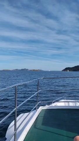 A boat is moving on the sea, creating a view of the ocean and the sky. The camera is placed on the deck of the boat, focusing on a railing in the foreground. In the distance, there are islands, coastline, and some buildings that appear to be historical structures. The sky is blue with scattered clouds, and the water is a deep blue. The movement of the boat creates a slight tilt in the video.