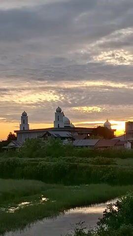 A time-lapse video shows a golden sunset over a city skyline with a mosque, houses, and green marshland with a small stream in the foreground. The sky is filled with clouds, and the sun sets behind the buildings.