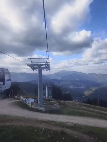 The video shows a ski lift tower and surrounding mountainous landscape under a cloudy sky. The ski lift tower is tall and metallic, with a cable running overhead. In the background, there are rolling hills and mountains covered in trees, with a few buildings visible in the distance. The ground in the foreground is covered in grass and dirt paths, with some wooden fencing. A person on a bicycle is visible riding down a winding path in the middle ground. The overall scene appears to be a scenic outdoor location, possibly a mountain resort.