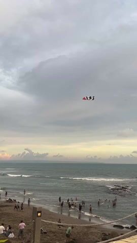 The video shows a beach scene on a cloudy day. There are people on the beach and in the water. A kite is flying in the sky. The sky is filled with grey clouds, and a hint of sunset is visible on the horizon. The ocean has small waves. The beach has dark sand. Some people are wading in the water, while others are on the sand, some sitting and some standing. The kite is black and white, with a small section of red.
