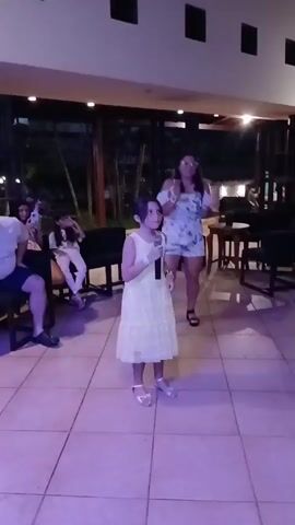 A young girl in a yellow dress and sandals stands center frame holding a microphone and singing. She has dark hair pulled back and a light-colored bracelet on her left wrist. Behind her, a woman in a floral romper and glasses stands clapping. To the left of the young girl, a man with a beard and glasses sits on a dark chair, looking at his phone. Beside him, a girl in white pants and a patterned top leans back in her seat, looking up. The room has a tiled floor and the lighting is dim, with pinkish purple light on the floor in the foreground. The background shows furniture, including chairs and tables, and large windows looking out onto a dark, possibly outdoor, area with some greenery.