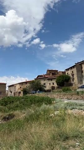 The video shows a hillside village under a blue sky with white clouds. The village is built on a slope, with stone buildings and red tile roofs. The foreground is filled with green grass and vegetation. The camera pans across the scene, revealing more of the village and the surrounding landscape.