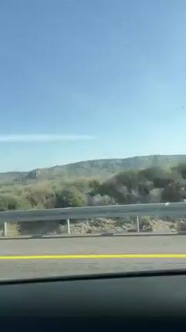 The video shows a view from a car driving on a road. The road has a metal guardrail and a yellow line marking. Beyond the guardrail, there's a landscape with green fields and some scrubby vegetation. In the distance, there are hills. The sky is blue and clear.