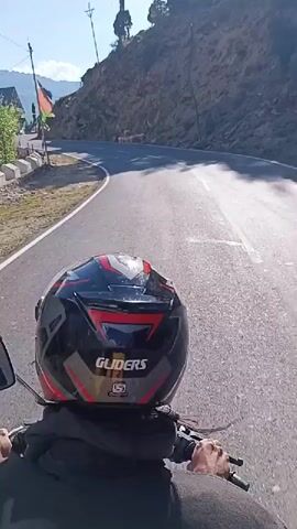The video shows a first-person perspective of someone riding a motorcycle on a paved road in a mountainous region. The rider is wearing a black and red helmet with "GLIDERS" written on the back. The road curves to the right, with a steep, rocky hillside on the right side and a drop-off to the left, where there are some buildings and trees. Several cars are visible on the road ahead, and the sky is clear and blue. The sun is shining, casting shadows on the road. There is some dust or smoke trailing behind the motorcycle.