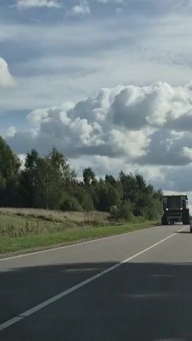 A combine harvester with a header attached is driving down a paved road on the right side of the frame. The combine is green and black, and the header is green and red. The road has white lane markings. To the left of the road is a grassy shoulder with trees and bushes. In the distance, there are large white clouds in a blue sky. A dark-colored car is visible on the road ahead of the combine. The video appears to be filmed from another vehicle moving alongside the combine.