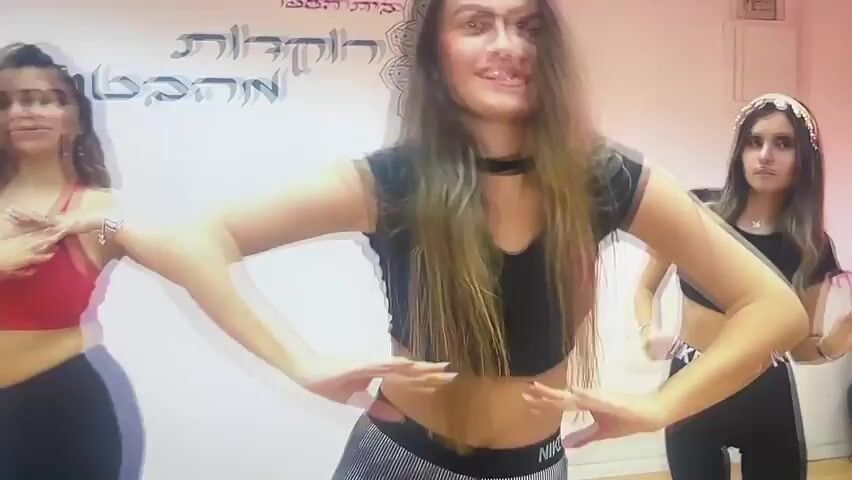 The video shows three young women in a dance studio. The woman in the center, with long, blonde-streaked hair, is wearing a black crop top and patterned leggings. She is smiling and appears to be leading a dance. To her left, another woman in a red sports bra and black leggings is also dancing. To the right, a third woman in a black crop top and black leggings with the "NIKE PRO" logo is dancing. The background features a white wall with some script in what appears to be Hebrew, and a stylized image of a dancer. The lighting suggests an indoor setting, likely during the daytime. The mood seems energetic and positive, with the women engaged in a dance routine. There is no indication of the time of day or weather conditions outside. The location is described as Shakhovskaya, Russia, but there are no visible cues in the video to confirm this.