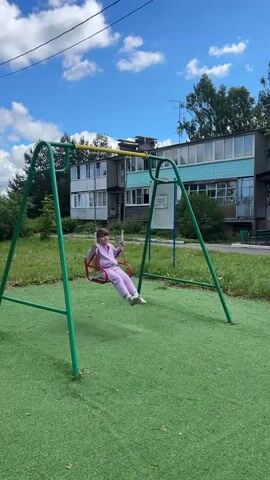 A young girl in a light purple tracksuit is joyfully swinging on a red swing set. The swing set has green metal A-frame supports and a yellow horizontal bar from which the swing is suspended by chains. The girl is in mid-air, her legs extended forward, suggesting she is enjoying a high swing.

The scene appears to be a small playground with a bright green, artificial turf surface. In the background, there is a patch of dry grass and some trees. Behind the trees, a multi-story apartment building with light blue and white siding is visible. The building has several windows and balconies. The sky is a vibrant blue with scattered white clouds, indicating a sunny day. A telephone pole with wires is visible to the left of the swing set.

The overall atmosphere is one of childhood play and happiness. The girl's posture and the dynamic movement of the swing suggest a moment of exhilaration. The setting appears to be an urban or suburban residential area, likely in Russia, given the mention of "Борщевка Малая." There is no visible text in the frame. The time of day is daytime, as evidenced by the bright sunlight and shadows.