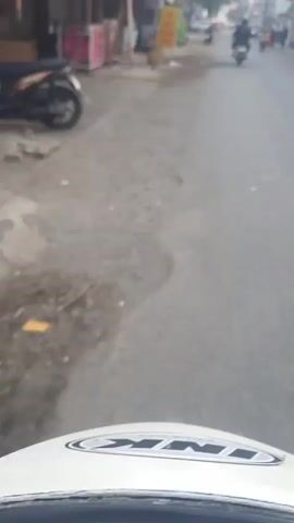 The video shows a slightly blurry, street-level view from what appears to be a moving vehicle, possibly a motorcycle, looking forward down a paved road. The road is flanked by buildings on both sides, suggesting a commercial or residential area. On the left side of the road, there are buildings with signs and some debris. On the right side, there are more buildings with shop fronts. Several people are visible walking and riding motorcycles on the road. Some individuals on the right side of the road are wearing light-colored shirts and helmets, and they appear to be walking together. The lighting suggests it is daytime, possibly early morning or late afternoon, as the sky is overcast. The overall impression is a bustling street scene in a town or city.