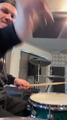 A young man wearing a black baseball cap and a black hoodie is playing drums in a room with acoustic paneling. He is smiling and sticking his tongue out as he plays. He's focusing on a snare drum and cymbals. The room is brightly lit, with a white ceiling.