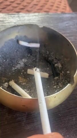 A close-up shot shows a hand holding a cigarette over an ashtray filled with cigarette butts and ash. The ashtray appears to be made of brass and is quite full, indicating multiple cigarettes have been smoked recently. One cigarette butt is positioned directly in the center of the ashtray, and another is actively being held by a person's fingers. The background shows a textured surface, possibly a table or placemat. The lighting is natural and appears to be from overhead. The overall impression is one of casual smoking in an indoor setting.
