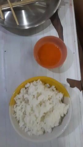 A top-down shot of a meal prepared for eating. In the foreground, there is a white rice in a yellow bowl, and a small orange bowl with orange sauce. Behind these is a large stainless steel pot with chopsticks resting on it. Further back, some food is being cooked in a black pan, with pieces of meat and onion. There are also two clear plastic containers with more food. The scene is brightly lit and appears to be indoors.