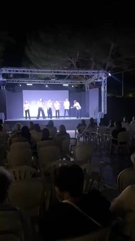 A group of six individuals, five on stage and one in the audience, are present in the video. The five individuals on stage are positioned at different points, and appear to be performers engaged in some form of theatrical production. The audience member is seated and facing the stage. The environment is an outdoor setting at night, with a stage, lighting equipment, and trees visible. The stage features a screen behind the performers, and the overall lighting suggests an evening event. The audience is seated in chairs arranged in rows, facing the stage.