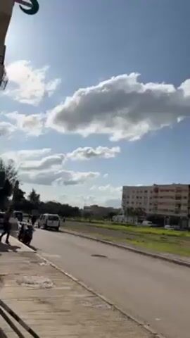 The video shows a scene on a sunny day with a partly cloudy sky. A road runs along a sidewalk with green space. A few people are present in the vicinity. Several cars are parked along the side of the road, including a gray pickup truck with items in its bed. A building with a colorful patterned facade is visible in the background.  The camera appears to pan slowly to the right, focusing on the parked vehicles and the nearby trees.