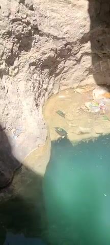 The video shows a deep, narrow gorge with light-colored, rough rock walls. A small stream of water trickles down one wall into a pool of turquoise water.  Some plastic trash is visible in the pool. The camera tilts upward to show the sky at the top of the gorge.