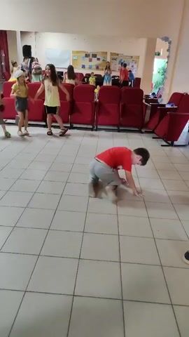 A boy in a red shirt and grey pants is breakdancing on a tiled floor in front of an audience of children and adults. He is performing a move where he balances on his hands with his legs in the air. The audience is seated in red chairs in a room that appears to be a community center or school auditorium. There are posters on the wall behind the audience.