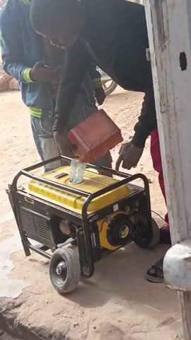 A person is pouring liquid from a red container into the gas tank of a yellow generator. Another person in a blue jacket is standing nearby, looking at a phone. The generator is on wheels and is sitting on a dirt surface. The camera then pans away and focuses on a brown surface with a bracelet with pink and white beads and a white cloth.