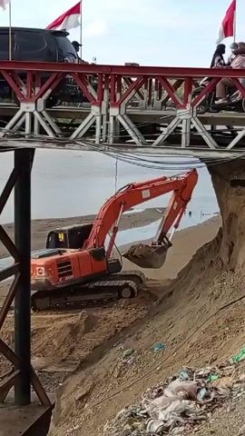 The scene shows an orange HITACHI excavator working on a sandy area adjacent to a river, likely performing maintenance or construction activities. The excavator is positioned near a dirt embankment and is actively digging into the sand. A bridge spans above the river and the excavator. Several motorbikes with people are crossing the bridge. Three Indonesian flags are flying atop the bridge, suggesting a local context or event. The environment includes a river, a sandy bank, and a bridge structure. The debris is scattered near the embankment, indicating ongoing construction or cleanup work.