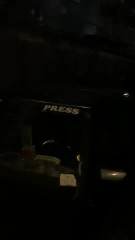 The image captures a dark vehicle, possibly a car or SUV, parked outdoors at night. The word "PRESS" is clearly visible in white capital letters on the side of the vehicle, suggesting its purpose or affiliation. In the dimly lit background, architectural structures, including buildings with windows and what appears to be a fence or gate, are partially visible, indicating an urban or suburban setting. The extreme darkness of the scene confirms it is nighttime, with only faint ambient light illuminating the surroundings. No people are visible, and no specific activity is occurring. A small, lighter-colored object or sticker is also visible on the lower side of the vehicle.