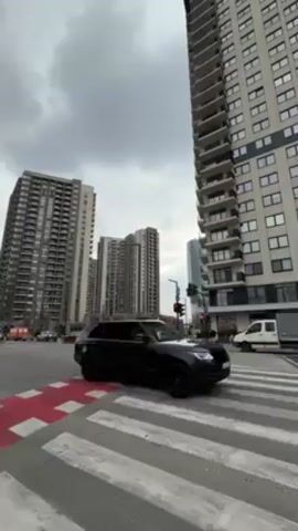 The camera is at street level, looking up at tall apartment buildings on a cloudy day. There is a crosswalk with red and white stripes, and cars are passing through the intersection. The traffic light is red. The cars include an SUV, a black hatchback, a white sedan, and a red tractor.