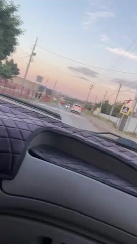 The video shows a view from inside a vehicle looking out through the windshield. In the foreground, the padded dashboard of the vehicle is visible. The road ahead is paved, and there is a pedestrian crossing marked with white stripes. Two white cars are visible on the road, one further ahead and one slightly closer. The sky is a soft gradient of pink, orange, and blue, indicating either sunrise or sunset. There are utility poles and wires along the side of the road. Buildings and fences are visible in the background. A yellow pedestrian crossing sign is also visible on the right side of the road. The overall scene suggests a quiet drive in a semi-urban or rural setting during twilight.