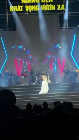 A female singer in a flowing white dress performs on a brightly lit stage with a band during a nighttime concert in Măng Đen, Vietnam. The stage features multiple tiers, neon pink and blue geometric light structures, and two large reflective or projected disco balls on either side. Numerous spotlights illuminate the stage and performers. Behind the band, a large screen or backdrop displays the text "MANG ĐEN KHÁT VỌNG VƯƠN XA" (Măng Đen - Aspiring to Reach Far). The singer is holding a microphone and gesturing, while the band members (drummer, guitarists, keyboardist) play their instruments. A large audience, visible as silhouettes in the foreground, watches the performance.