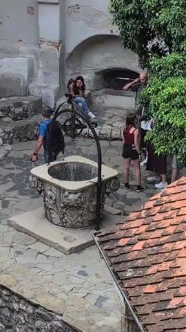 A group of people are gathered around an old well in a courtyard. The well has an ornate stone base and a metal crank mechanism. A man in a white shirt and shorts is leaning over the well, appearing to drink from it. A woman in a black dress is standing to the left of the well, looking towards the man. Several other people are observing the scene. The courtyard is paved with stones and surrounded by old stone buildings and trees.