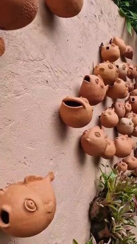 The video shows a wall decorated with numerous small, terracotta fish-shaped ornaments. The ornaments are of varying sizes and appear to be handcrafted, with some exhibiting more detailed features than others. They are attached to a textured, light-colored wall. Some of the ornaments have open mouths, giving the impression of them swimming or blowing bubbles. In the lower right corner of the frame, there are some green plants in pots, suggesting an outdoor or garden setting. The lighting is natural, casting soft shadows.