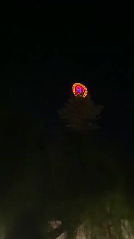 A nighttime scene unfolds with a large, brightly lit, circular object visible at the top of a tree. The object has concentric rings of red and blue light. The camera then pans down to reveal a crowd of people gathered outdoors at night. The crowd is facing a stage area with a structure that appears to be a lighting and sound rig. Buildings with illuminated windows are visible in the background. The lighting suggests an outdoor event or festival.
