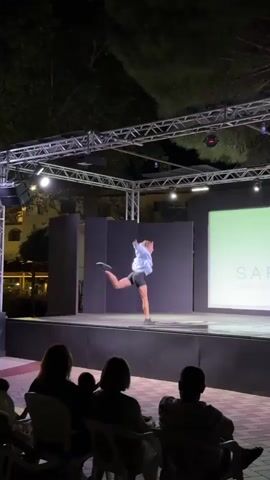 A female dancer performs on an outdoor stage at night. She is wearing a light blue shirt and dark shorts. The stage is lit with overhead lights and a large screen is behind her showing a logo. An audience of about five people is seated in front of the stage.